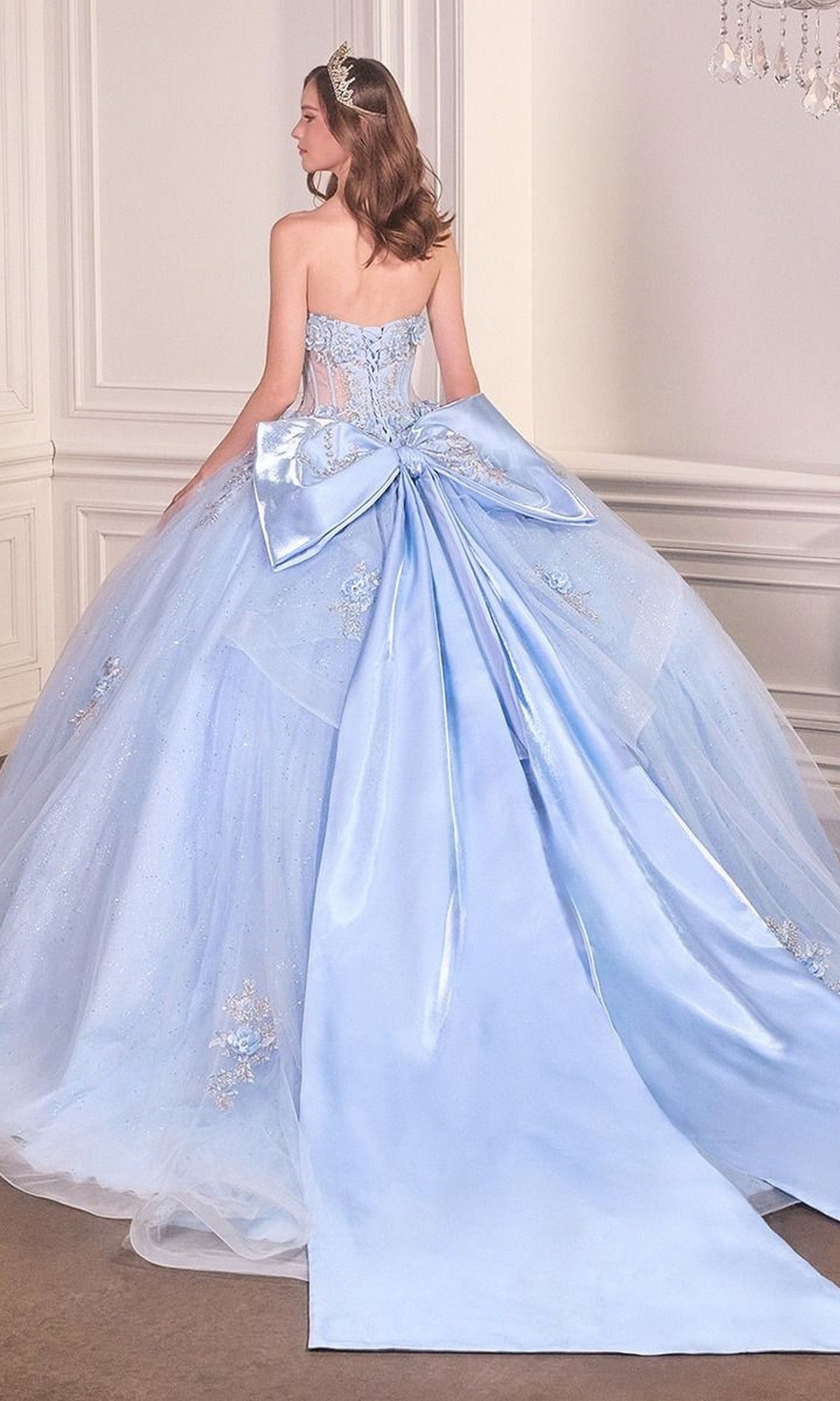 A woman stands in an elegant room, facing away in the Quinceanera Dress 15726 By Ladivine—a strapless, light blue ball gown featuring a large bow, glitter tulle, and a long train.